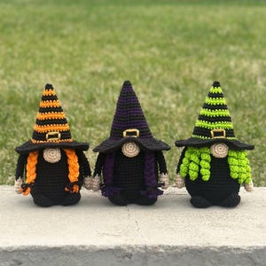 May include: Three crocheted gnome figures wearing pointed hats with gold-colored buckles. The left gnome has orange and black striped hat with orange braids. The center gnome has a purple and black hat with purple braids. The right gnome has a lime green and black striped hat with lime green curls.