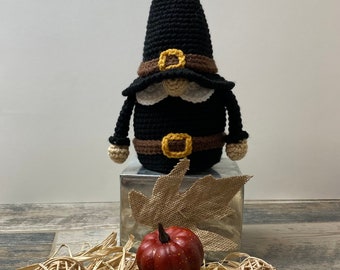 Pode incluir: Um gnomo de Ação de Graças de crochê com um chapéu preto, um cinto marrom e amarelo e uma barba branca. O gnomo está em pé em uma caixa prateada com uma folha de juta e uma pequena abóbora vermelha.