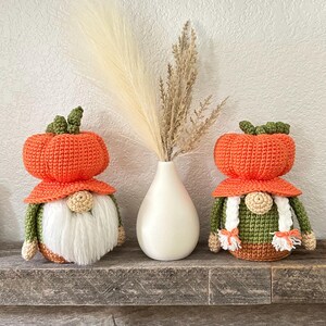 Two handmade crochet gnomes with orange pumpkin-shaped hats and green bodies, positioned on either side of a white teardrop-shaped vase containing dried beige pampas grass. The left gnome has a white beard, and the right has white braids.