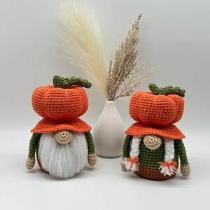 Two handmade crochet gnomes featuring orange pumpkin-shaped hats with green stems. The left gnome has a long white beard, while the right gnome has two white braided pigtails with orange ties. Both wear green and brown bodies with tan hands. Digital PDF pattern.