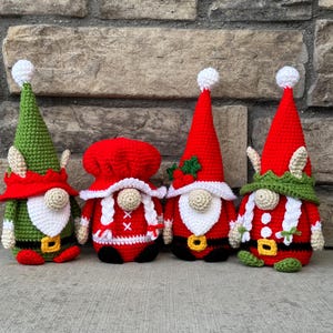 Four crochet Christmas gnomes posed in front of a stone wall, including Santa, Mrs. Claus, and two elf gnomes in red, green, and white festive outfits with tall hats and belts, handmade amigurumi figures shown for the downloadable PDF crochet pattern.