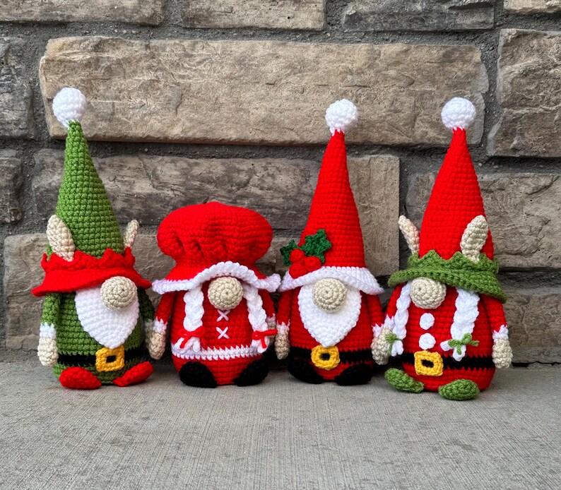 Four crochet Christmas gnomes posed in front of a stone wall, including Santa, Mrs. Claus, and two elf gnomes in red, green, and white festive outfits with tall hats and belts, handmade amigurumi figures shown for the downloadable PDF crochet pattern.