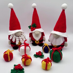 Three crochet Christmas gnomes with tall red hats surrounded by colorful crocheted gift boxes and holly decorations, handmade amigurumi figures in festive red, white, and green colors shown for the downloadable PDF crochet pattern.