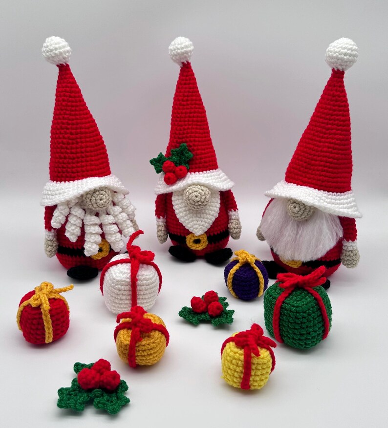 Three crochet Christmas gnomes with tall red hats surrounded by colorful crocheted gift boxes and holly decorations, handmade amigurumi figures in festive red, white, and green colors shown for the downloadable PDF crochet pattern.