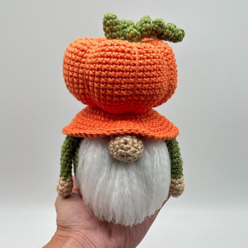 A crochet gnome featuring a large orange pumpkin-shaped hat with a green stem top. The gnome has a round beige nose, white fluffy beard, and a dark green body with a brown base.