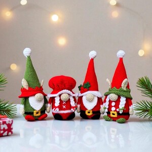 Four festive crochet Christmas gnomes posed on a white surface with soft twinkling holiday lights and pine branches in the background, including Santa, Mrs. Claus, and two elf gnomes in red, green, and white outfits, handmade amigurumi figures featured for the downloadable PDF crochet pattern.