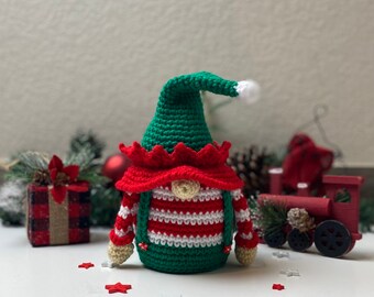 May include: A handmade crochet elf doll with a green hat, red and white striped shirt, and green suspenders. The elf has a red collar and a small beige nose. The doll is decorated with red and white stars, and a small gift box.