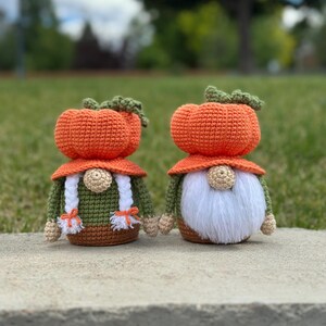 Two handmade crochet gnomes featuring orange pumpkin-shaped hats with green stems. The left gnome has white braided hair with orange ties, while the right gnome has a long white beard. Both wear olive green shirts and brown bases. Digital PDF pattern.