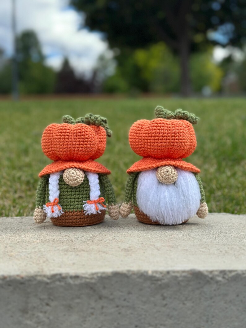 Two handmade crochet gnomes featuring orange pumpkin-shaped hats with green stems. The left gnome has white braided hair with orange ties, while the right gnome has a long white beard. Both wear olive green shirts and brown bases. Digital PDF pattern.