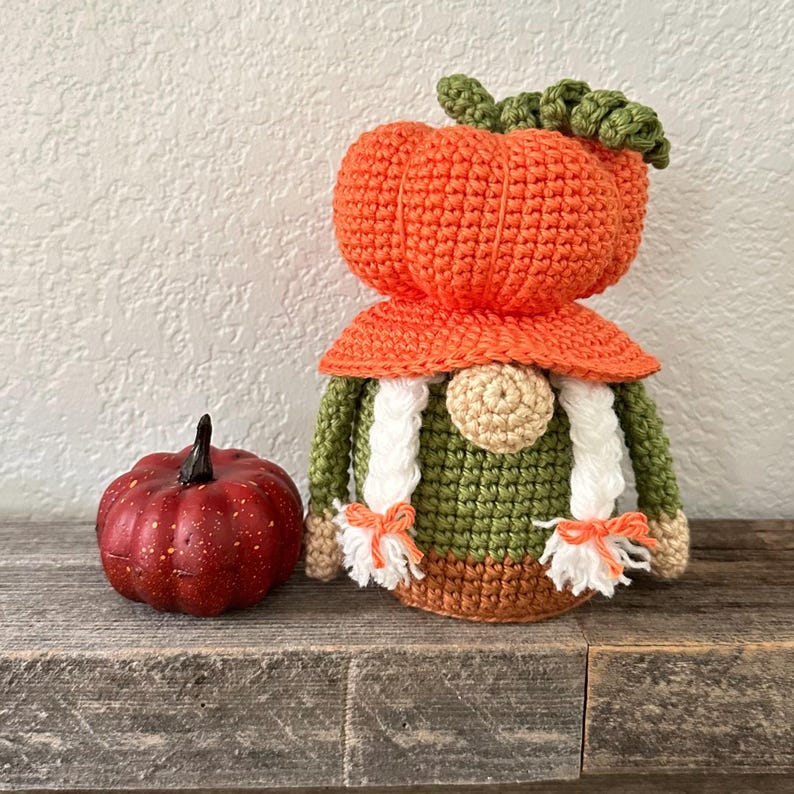 A crochet gnome figure featuring a large orange pumpkin-shaped hat with a green stem, a round beige nose, and two white braided pigtails with orange ties, positioned next to a small, dark red, textured decorative pumpkin.