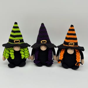 Può includere: Tre gnomi all'uncinetto che indossano cappelli da strega a punta con fibbie color oro. Il primo gnomo ha un cappello a righe verde lime e nero con riccioli verde lime. Il secondo ha un cappello viola e nero con trecce viola. Il terzo ha righe arancioni e nere con trecce arancioni.