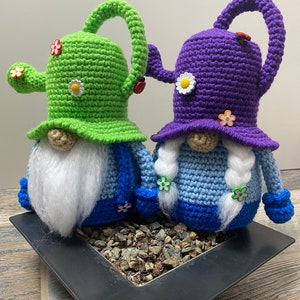 May include: Two crocheted gnomes wearing watering can hats. The gnome on the left is wearing a green watering can hat and the gnome on the right is wearing a purple watering can hat. Both gnomes are wearing blue and white sweaters and have white beards.