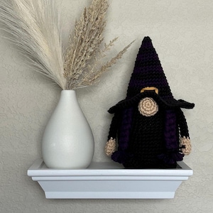 A crocheted Halloween witch gnome wearing a tall, pointed hat with horizontal black and purple stripes, a wide black brim, a tan nose, and purple braided hair, positioned next to a white vase filled with dried pampas grass. This is a downloadable crochet pattern PDF.