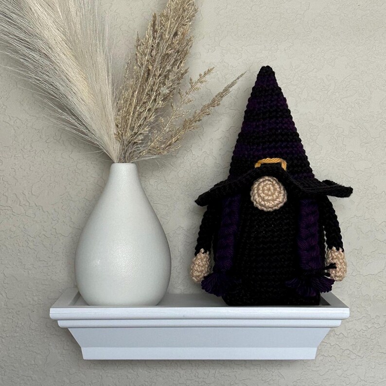 A crocheted Halloween witch gnome wearing a tall, pointed hat with horizontal black and purple stripes, a wide black brim, a tan nose, and purple braided hair, positioned next to a white vase filled with dried pampas grass. This is a downloadable crochet pattern PDF.