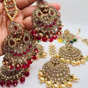 May include: A set of four gold-tone Indian bridal jewelry pieces with red and white stones. The pieces include a pair of earrings, a nose ring, and a forehead ornament.
