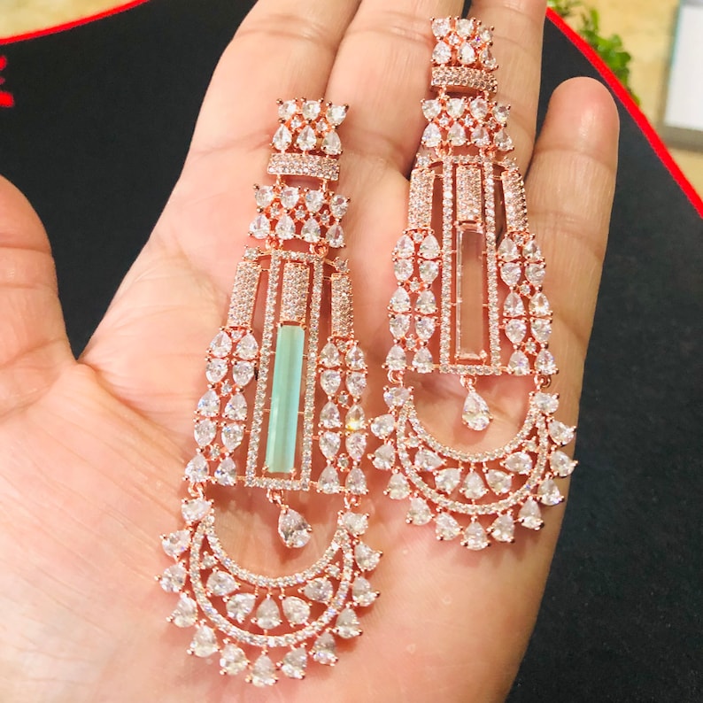 Puede incluir: Un par de pendientes largos de oro rosa con un dise&ntilde;o geom&eacute;trico. Los pendientes est&aacute;n adornados con piedras de circonita c&uacute;bica transparentes y una piedra rectangular azul claro en el centro.
