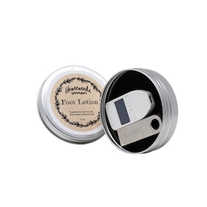 Puede incluir: Una lata de plata con una etiqueta que dice "Homemade Foot Lotion" y una lista de ingredientes. La lata está abierta y dentro hay una billetera de hardware de criptomonedas Ledger Nano S plateada.