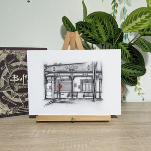 May include: A black and white drawing of a shop front with the word "MAGIC" above the entrance. The drawing is on a white background and is framed on a wooden easel.