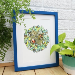 May include: A framed watercolour painting of a circular arrangement of autumn leaves and acorns. The leaves are green, yellow, and brown, and the acorns are brown with a hint of blue. The background is a pale yellow.