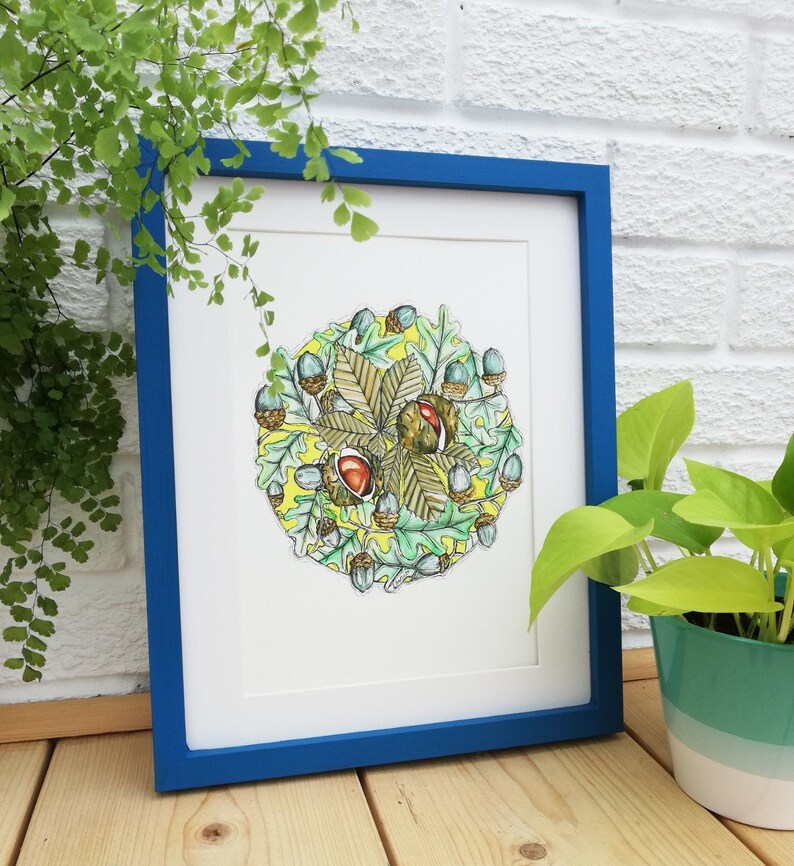 May include: A framed watercolour painting of a circular arrangement of autumn leaves and acorns. The leaves are green, yellow, and brown, and the acorns are brown with a hint of blue. The background is a pale yellow.