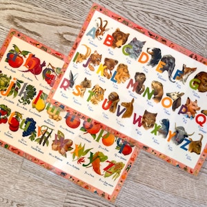 May include: Two vintage alphabet posters. One features animals, the other fruits and vegetables. Each poster has a decorative pink border. The letters are in a colourful font with corresponding illustrations. The posters are on a light wood surface.