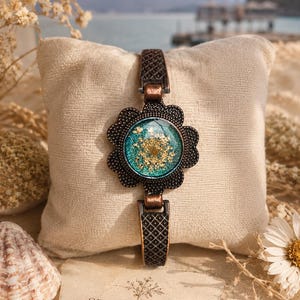 May include: A floral-themed bracelet with a flower-shaped centerpiece. The centerpiece has a teal background with dried flowers encased in resin. The bracelet has a dark bronze finish with a patterned band. The card reads "SENORITA GAIA Nature's Touch, Handmade for You."