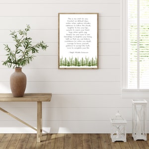 May include: A framed print with a wooden frame displays a poem by Ralph Waldo Emerson. The text is in black font on a white background, with a watercolor forest design at the bottom. The print is displayed on a wooden table with a vase of greenery.
