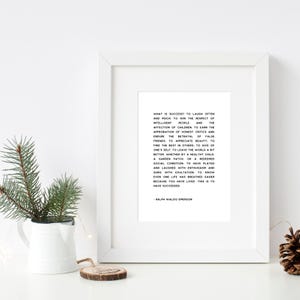 May include: A framed white print with black text quoting Ralph Waldo Emerson on success. The print is displayed on a white surface, with a small white pitcher holding greenery and a pine cone to the side.