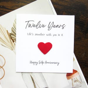 K&ouml;nnte beinhalten: Eine wei&szlig;e Karte mit den Worten "Twelve Years" und "Life's smoother with you in it" in schwarzer Schrift. Ein rotes Herz ist auf der Karte befestigt. Die Worte "Happy Silk Anniversary" sind darunter gedruckt. Die Karte liegt auf einer wei&szlig;en und beigen Oberfl&auml;che.