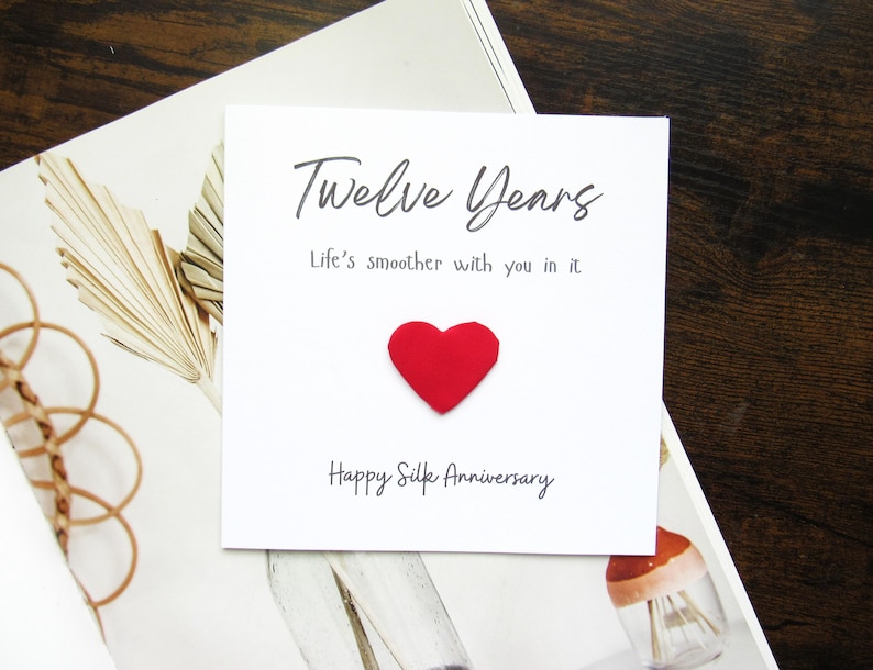 K&ouml;nnte beinhalten: Eine wei&szlig;e Karte mit den Worten "Twelve Years" und "Life's smoother with you in it" in schwarzer Schrift. Ein rotes Herz ist auf der Karte befestigt. Die Worte "Happy Silk Anniversary" sind darunter gedruckt. Die Karte liegt auf einer wei&szlig;en und beigen Oberfl&auml;che.