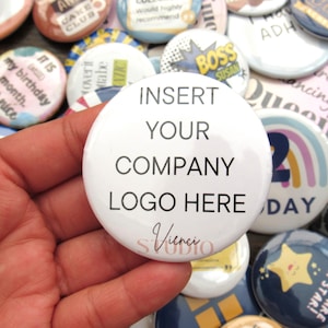 Op de afbeelding: Een verzameling ronde, witte buttons met diverse ontwerpen en tekst. Eén button op de voorgrond leest "INSERT YOUR COMPANY LOGO HERE". Andere buttons bevatten zinnen als "my birthday month" en "BOSS SUSAN."