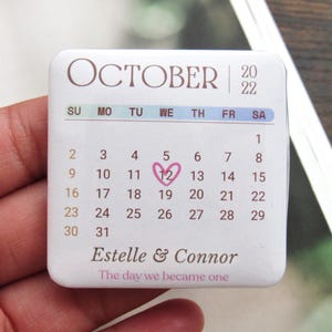 Puede incluir: Un imán de calendario cuadrado blanco para octubre de 2022, con el número 12 circulado en rosa. El calendario incluye los nombres Estelle & Connor y el texto "The day we became one."