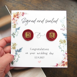 May include: A white wedding card with floral borders, featuring the text "Signed and sealed." Two red wax seals with gold initials are displayed. The card also includes the text "Congratulations on your wedding day 12.9.2027."