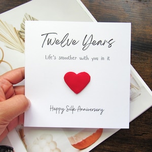 K&ouml;nnte beinhalten: Eine wei&szlig;e Karte mit dem Text "Twelve Years" und "Life's smoother with you in it." Ein rotes Herz ist auf der Karte befestigt, mit den Worten "Happy Silk Anniversary" darunter. Die Karte wird &uuml;ber einem dekorativen Hintergrund gehalten.
