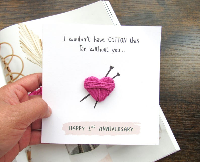 May include: A white anniversary card featuring a pink yarn heart with two black knitting needles. The card has the text "I wouldn't have COTTON this far without you..." and "HAPPY 2ND ANNIVERSARY". The card rests on a magazine.