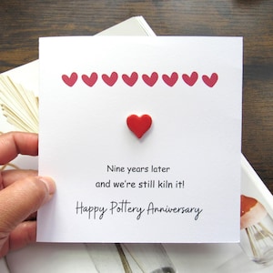 Könnte beinhalten: Weiße Karte mit rotem Herz und neun kleineren roten Herzen. Der Text lautet: "Nine years later and we're still kiln it! Happy Pottery Anniversary."