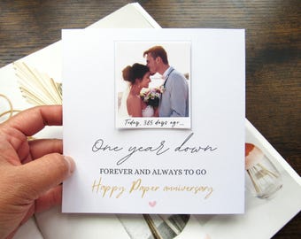 May include: A white anniversary card with a photo of a couple. The card reads "One year down, forever and always to go, Happy Paper anniversary." A small pink heart is at the bottom. The photo is of a couple kissing.