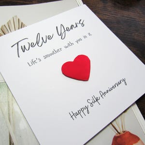 K&ouml;nnte beinhalten: Eine wei&szlig;e Gru&szlig;karte mit den Worten "Twelve Years" und "Life's smoother with you in it" in schwarzer Schrift. Ein erhabenes rotes Herz befindet sich in der Mitte der Karte. Darunter steht "Happy Silk Anniversary".