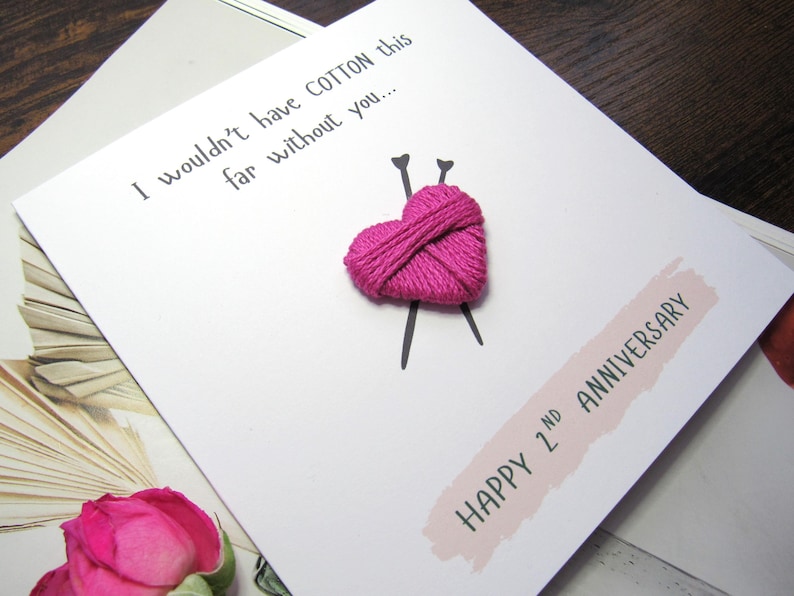 May include: White card featuring a pink embroidered heart and knitting needles. The text reads "I wouldn't have COTTON this far without you..." and "HAPPY 2nd ANNIVERSARY" on a pink banner. A pink rose is visible.