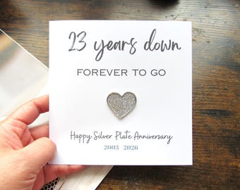 May include: A white card with the text "23 years down FOREVER TO GO" and "Happy Silver Plate Anniversary 2003 2026". A silver glitter heart is affixed to the card. The card is held by a hand.