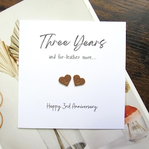 May include: A white card with the text "Three Years and for-leather more..." and "Happy 3rd Anniversary" in black script. Two brown heart-shaped embellishments are below the text. The card is on a white and brown surface.