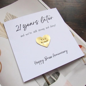 May include: A white card with the text "21 years later" and "Happy Brass Anniversary". A gold heart-shaped pin is attached. The card rests on a patterned background.