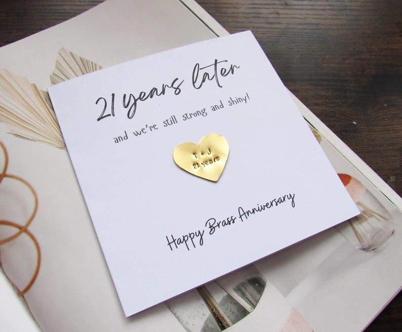 May include: A white card with the text "21 years later" and "Happy Brass Anniversary". A gold heart-shaped pin is attached. The card rests on a patterned background.