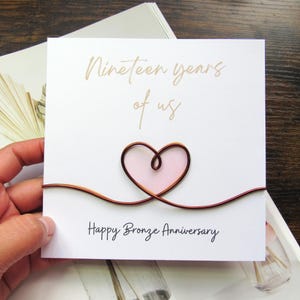 以下が含まれることがあります： 「Nineteen years of us」の金色の筆記体で書かれた白いカード。ピンク色の中心を持つ銅線で作られたハートが中心です。「Happy Bronze Anniversary」の文字が下に印刷されています。