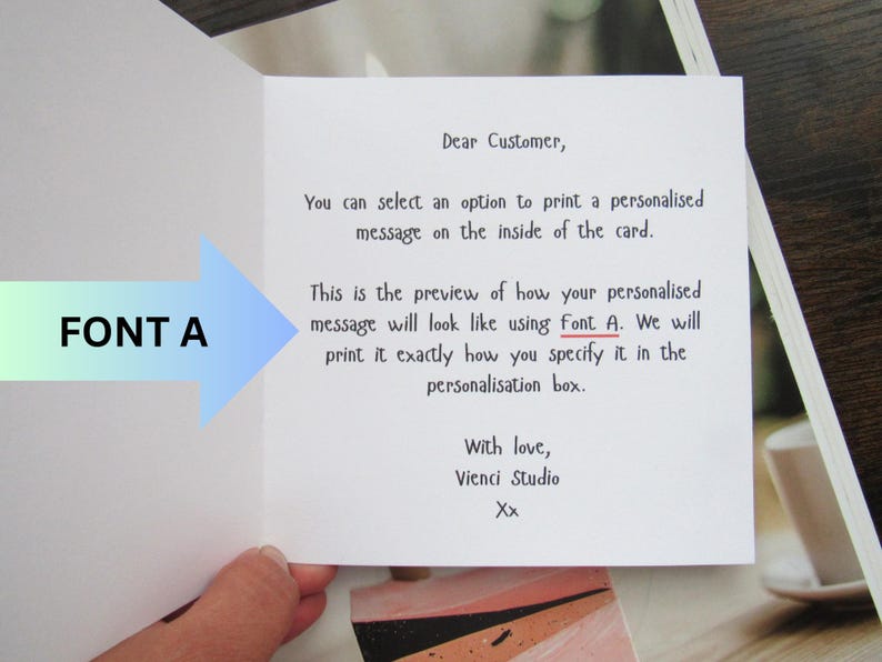 May include: An open greeting card with text explaining how a personalised message will appear using Font A. The card is white with black text and a blue and green arrow pointing to the text.