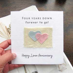 Puede incluir: Una tarjeta blanca con el texto "Four Years Down forever to go!" y "Happy Linen Anniversary". Presenta un cuadrado de tela beige con una aplicación de corazón azul y rosa. La tarjeta está sujeta por una mano.