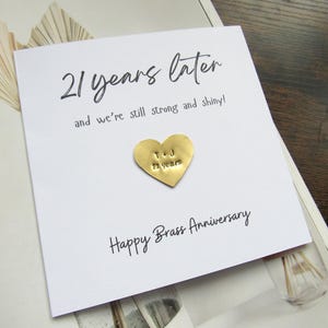 May include: A white card with the text "21 years later" and "Happy Brass Anniversary". A gold heart-shaped charm is attached, engraved with the initials "T & J" and "21 years".