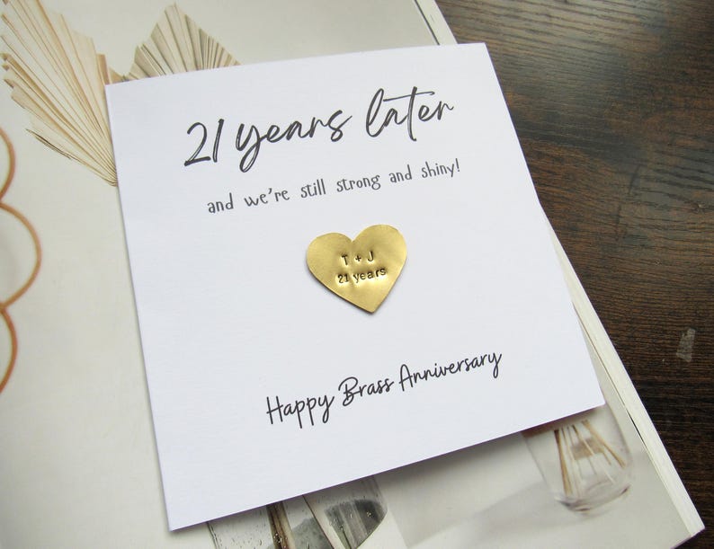 May include: A white card with the text "21 years later" and "Happy Brass Anniversary". A gold heart-shaped charm is attached, engraved with the initials "T & J" and "21 years".