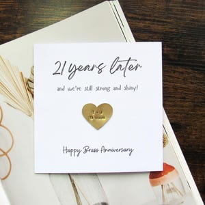 May include: A white card with the text "21 years later" and "Happy Brass Anniversary". A small gold heart with "T & J 21 years" is affixed to the card. The card is on a magazine.