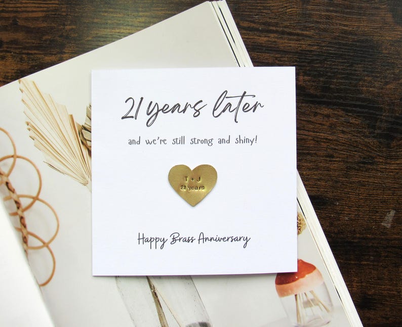 May include: A white card with the text "21 years later" and "Happy Brass Anniversary". A small gold heart with "T & J 21 years" is affixed to the card. The card is on a magazine.